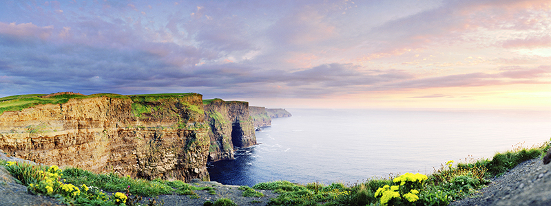 Bruna klippor med blommor och grönska vid ett öppet hav, Cliffs of Moher på Irland.