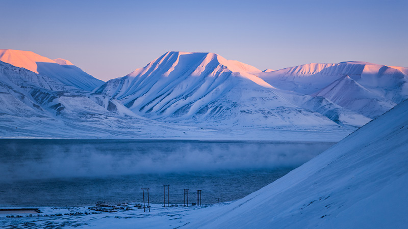 Gryning på Svalbard, snöklädda berg och en stillsam sjö.